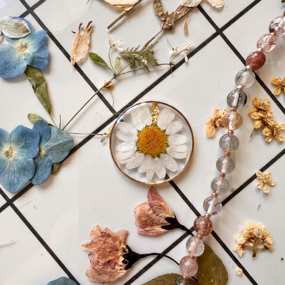 Colgante de resina transparente con una margarita natural, rodeado de flores prensadas para el taller de joyería botánica